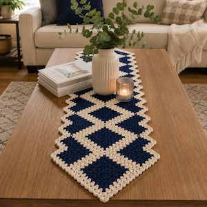 May include: A rectangular crochet table runner featuring a geometric diamond pattern in navy blue and cream yarn. The runner is centered on a wooden table, topped with a white ribbed ceramic vase containing green eucalyptus branches and a glass candle.