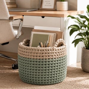 May include: A two-tone, round, crochet storage basket in sage green and beige. The basket is filled with notebooks, pens, and pencils. The basket has two handles and sits on a neutral-colored rug near a desk and potted plant.