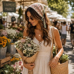 Può includere: Un foulard all'uncinetto con un motivo a piastrelle granny multicolore nei toni del rosa, verde e crema, annodato sul retro. Il foulard è indossato con un abito bianco senza maniche con dettagli in pizzo e una borsa in paglia intrecciata.