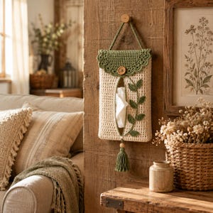 May include: A cream-coloured crochet tissue holder with a green scalloped flap, a wooden button closure, and a green vine appliqué. The holder features a central vertical opening for tissues, a hanging loop, and a green tassel at the bottom.