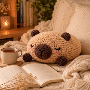 May include: A tan crochet plush toy shaped like a capybara with a dark brown snout, ears, and feet. The plush features embroidered closed eyes and pink cheeks. It is positioned on a textured surface alongside an open book and a ceramic mug.