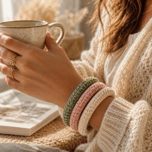 Può includere: Tre braccialetti all'uncinetto in verde oliva, rosa chiaro e crema. I braccialetti sono impilati su un polso. I braccialetti sono spessi e hanno un aspetto strutturato. Una persona tiene in mano una tazza di caffè.
