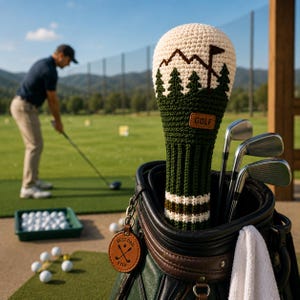 May include: A crochet golf club head cover featuring a cream top with a dark green mountain and pine tree design, a dark green body with a brown and cream striped base, and a brown leather tag labelled GOLF and a round tag reading BEST DAD EVER.
