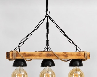 Puede incluir: Una lámpara colgante de tres luces con una viga de madera horizontal suspendida por cadenas de metal negro. Tres pantallas de cúpula de metal negro albergan bombillas redondas estilo Edison. La viga está envuelta con cuerda.