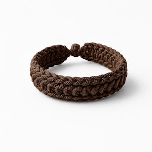 Può includere: Un braccialetto marrone scuro realizzato con una spessa corda intrecciata. L'accessorio circolare presenta un motivo intrecciato e testurizzato lungo tutta la sua lunghezza e include un piccolo meccanismo di chiusura a nodo sulla parte superiore.