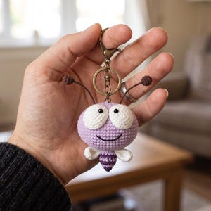 May include: A crochet keyring featuring a round, light purple creature with large white eyes, a small stitched smile, two antennae with dark brown tips, and a small striped purple and white body with two white wings attached to a bronze metal ring.