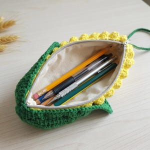 May include: A crochet pencil case shaped like a corn cob with a green husk exterior and yellow kernels. The case features a beige fabric lining, a silver zipper closure, and a green loop handle, containing four assorted pens and pencils inside.