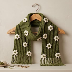 May include: An olive green ribbed knit scarf featuring white crocheted flower appliqués with dark green centers. The scarf has a fringed hem at both ends and is displayed on a wooden hanger. The design includes multiple floral accents.