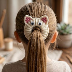 Könnte beinhalten: Ein cremefarbenes Häkel-Haargummi mit einem katzenartigen Design, zwei spitzen Ohren, rosa Details in den Ohren und zwei runden Wackelaugen. Das kreisförmige Accessoire hat eine mittige Öffnung und hält einen Pferdeschwanz aus hellbraunem Haar.