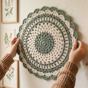Peut inclure: Un élément de décoration murale circulaire en crochet fait à la main, dans des tons de vert sauge et de crème. La pièce présente un bord festonné et un motif concentrique. L'objet est présenté sur un mur blanc, avec des impressions botaniques encadrées en arrière-plan.