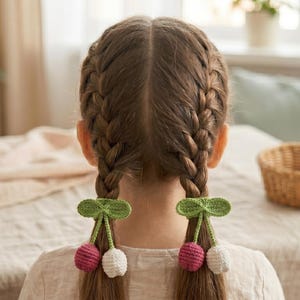 Puede incluir: Dos coleteros de ganchillo que presentan un par de cerezas cada uno, con hojas y tallos verdes unidos a una fruta esférica roja y otra blanca. Estos accesorios hechos a mano están sujetos alrededor de dos trenzas de pelo.
