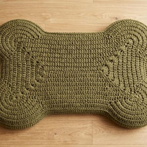 May include: A bone-shaped cushion featuring a textured olive green crochet knit pattern. The surface displays a consistent looped stitch design that follows the rounded contours of the four lobes and the central rectangular section of the object.