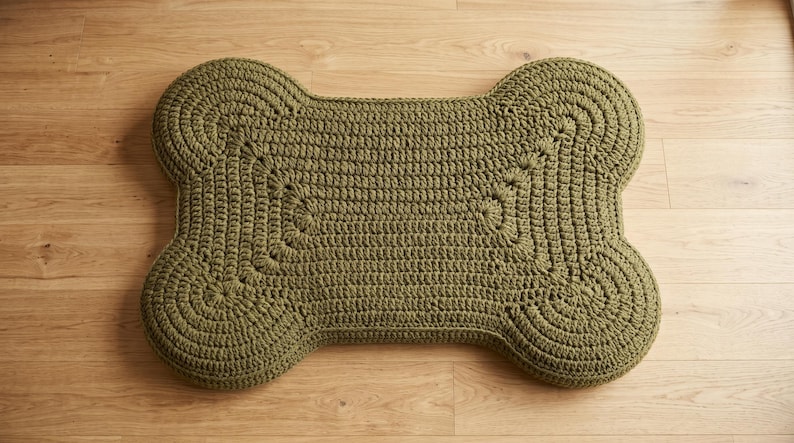 May include: A bone-shaped cushion featuring a textured olive green crochet knit pattern. The surface displays a consistent looped stitch design that follows the rounded contours of the four lobes and the central rectangular section of the object.