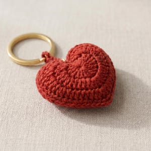 May include: A dark red, heart-shaped crochet keyring featuring a textured, yarn-stitched surface and a circular gold-toned metal split ring attached to the top. The heart is three-dimensional and crafted with visible, concentric circular stitching.
