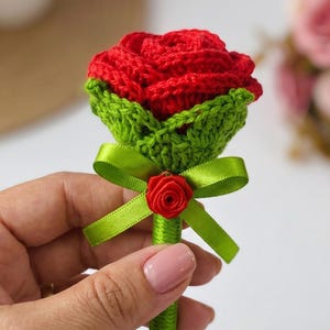May include: A crocheted red rose flower with green leaves attached to a stem wrapped in green ribbon. A small green satin ribbon bow is tied around the stem, featuring a tiny red fabric rose at the center of the bow knot.