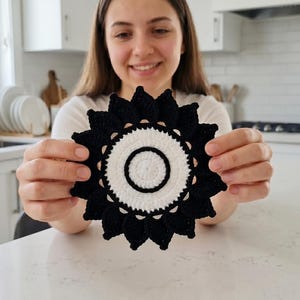 May include: A black and white crochet coaster with a sunburst design. The coaster is round with a central white circle and a black, pointed outer edge. The handmade item is held in front of a kitchen background.