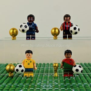 Könnte beinhalten: Miniaturfußballspieler-Figuren in verschiedenen Teamuniformen, die jeweils einen Fußball halten. Das Set enthält goldene Trophäen und den Text "Mini building block figurine". Die Figuren stehen auf einem grünen Feld.
