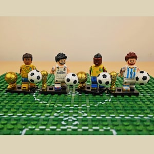 Könnte beinhalten: Vier Miniatur-Fußballspieler-Figuren mit Fußbällen und Trophäen auf einem grünen Feld. Die Spieler tragen verschiedenfarbige Trikots, darunter Gelb, Weiß und Blau. Jede Figur hält einen Fußball.