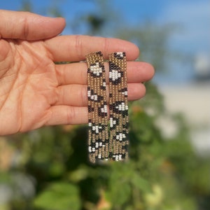 May include: A pair of long, beaded earrings with a black, white, and gold geometric pattern. The earrings are dangling and have a fringe-like appearance.