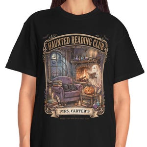 Op de afbeelding: Een zwart t-shirt met een grafische print van een gezellige kamer met een open haard, een geest die een boek leest, een pompoen en een kat. De tekst luidt HAUNTED READING CLUB EST 1892 en MRS. CARTER'S. Het ontwerp bevat kaarsen, boeken en een raam.