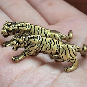 May include: Two small, metallic gold-coloured tiger figurines crafted with detailed black stripes and open mouths. Each tiger is depicted in a prowling stance with a curved tail, showcasing a textured finish that highlights the feline features.