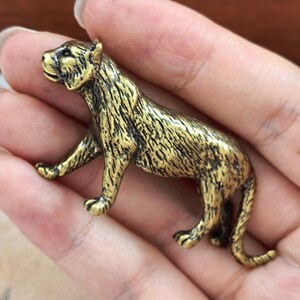 May include: A small, three-dimensional brass-coloured metal figurine of a tiger in a walking pose. The surface features a textured, antiqued finish with dark recessed areas highlighting the feline's muscular build, facial features, and tail.