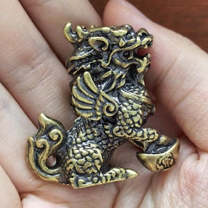 Op de afbeelding: Een klein, antiek ogend koperen beeldje van een mythisch wezen, waarschijnlijk een Pixiu of Qilin, met gedetailleerde schubben, vleugels en een gekrulde staart. Het wezen staat rechtop en houdt een kleine goudstaaf vast in zijn voorpoot.