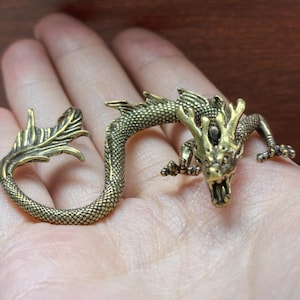 May include: A brass-colored metal dragon figurine with a textured, scale-like body, a long curved tail, and detailed facial features including horns and an open mouth. The dragon is depicted in a crawling position with its claws extended.