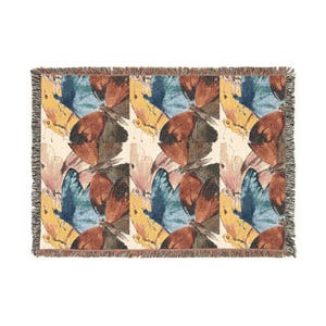 Puede incluir: Una manta tejida rectangular con un patrón repetitivo de mariposas en tonos naranja óxido, azul cerceta y amarillo mostaza sobre un fondo crema, terminada con un borde decorativo de flecos en los cuatro lados.