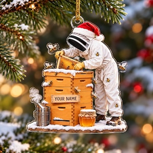 May include: A clear acrylic Christmas ornament featuring a beekeeper in a white suit and Santa hat tending to a wooden beehive covered in snow. The hive includes a sign reading YOUR NAME, a metal smoker, a jar of honey, and several bees.