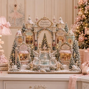 Op de afbeelding: Een gedetailleerd miniatuur kerstdorp genaamd Sophia's Ballet Village, met versierde pastelkleurige gebouwen, een centrale fontein, notenkrakerfiguren, miniatuur kerstbomen en zwaanbeelden, allemaal verlicht met warm interieurlicht.