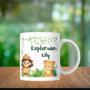 Puede incluir: Taza de cerámica blanca con una ilustración de dibujos animados de una niña exploradora y un cachorro de leopardo, con el texto "Exploradora Lily" en verde. La taza tiene un asa curva y está decorada con vides y hojas verdes.