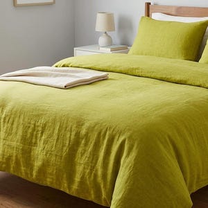 Puede incluir: Un juego de funda nórdica de color verde chartreuse con un acabado de tela texturizada, dos fundas de almohada a juego y una manta color crema doblada sobre la cama. La ropa de cama se muestra sobre una estructura de cama de madera.