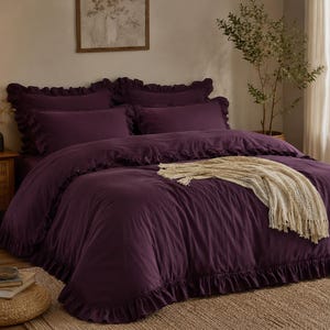 Könnte beinhalten: Ein tiefviolettes Bettwäsche-Set bestehend aus einem Bettbezug und passenden Kissenbezügen, die alle mit Rüschenkanten versehen sind. Eine cremefarbene Strickdecke mit Fransen liegt in der Mitte des Bettes und verleiht dem Set eine Textur.