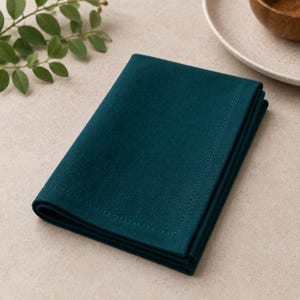 Puede incluir: Una servilleta de tela de color verde azulado oscuro plegada, con una textura lisa y mate y un detalle de borde cosido a lo largo de los lados. La tela está cuidadosamente doblada, mostrando un color profundo y sólido en toda la superficie.