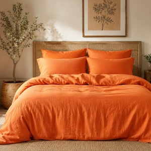 Puede incluir: Un juego de cama de color naranja vibrante que incluye una funda nórdica y cuatro almohadas rectangulares a juego. La tela presenta un tejido texturizado visible, creando una apariencia suave y relajada en toda la superficie de la cama.