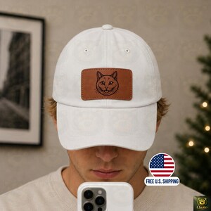 May include: A white baseball cap featuring a rectangular brown leather patch on the front. The patch is embossed with a black line drawing of a cat face. The cap has a curved brim and a structured crown with ventilation eyelets on the top panels.