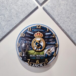 May include: A round wall clock featuring a Real Madrid theme with a stadium background, the club crest, a trophy, and the text "&iexcl;HALA MADRID!", "REAL MADRID 1902", and "JACK". The clock face has gold-colored numbers from 1 to 12 and black hands.