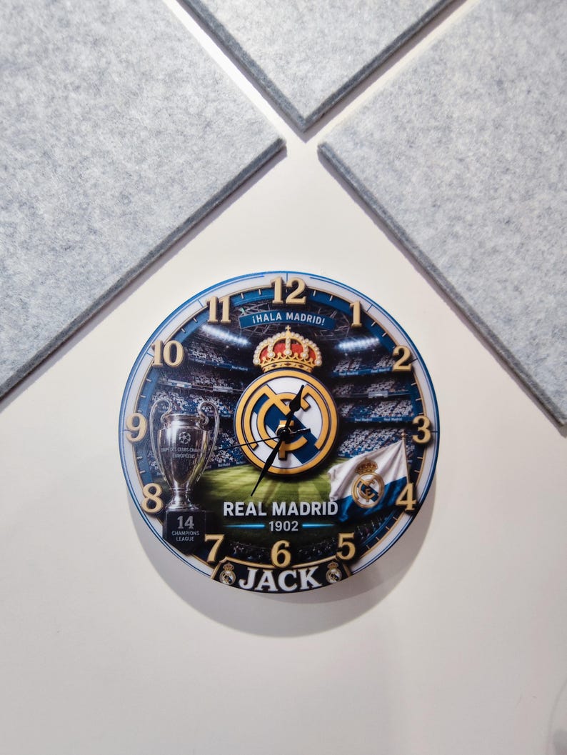 May include: A round wall clock featuring a Real Madrid theme with a stadium background, the club crest, a trophy, and the text "&iexcl;HALA MADRID!", "REAL MADRID 1902", and "JACK". The clock face has gold-colored numbers from 1 to 12 and black hands.