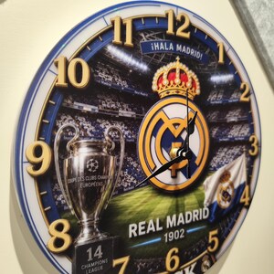 May include: A round wall clock featuring a Real Madrid theme with a stadium background, the club crest, a Champions League trophy, and the text "&iexcl;HALA MADRID!", "REAL MADRID 1902", "14 CHAMPIONS LEAGUE", and "JACK". It has gold-colored numbers and black hands.