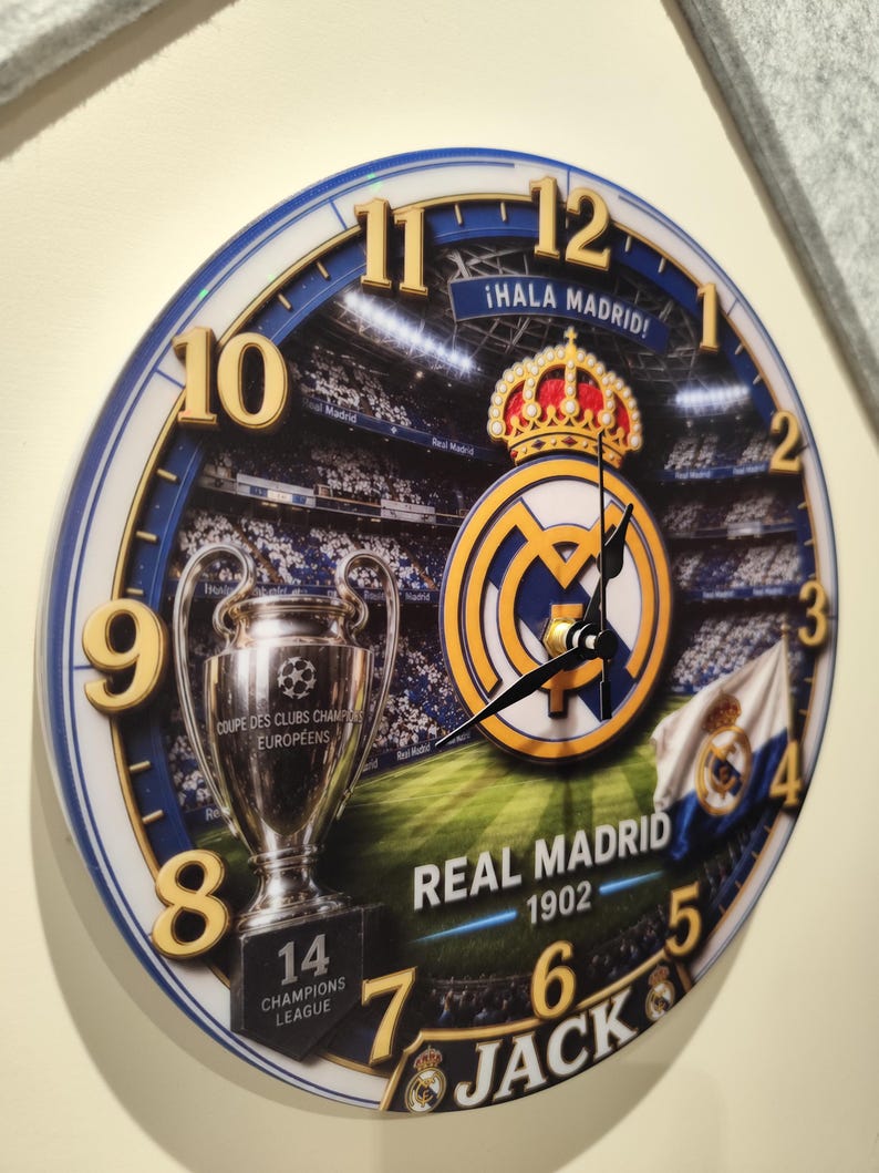 May include: A round wall clock featuring a Real Madrid theme with a stadium background, the club crest, a Champions League trophy, and the text "&iexcl;HALA MADRID!", "REAL MADRID 1902", "14 CHAMPIONS LEAGUE", and "JACK". It has gold-colored numbers and black hands.