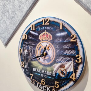 May include: A round wall clock featuring a Real Madrid theme with a stadium background, the club crest, a trophy, and a flag. The clock face displays the text "&iexcl;HALA MADRID!", "REAL MADRID 1902", and the name "JACK" at the bottom with gold numbers.
