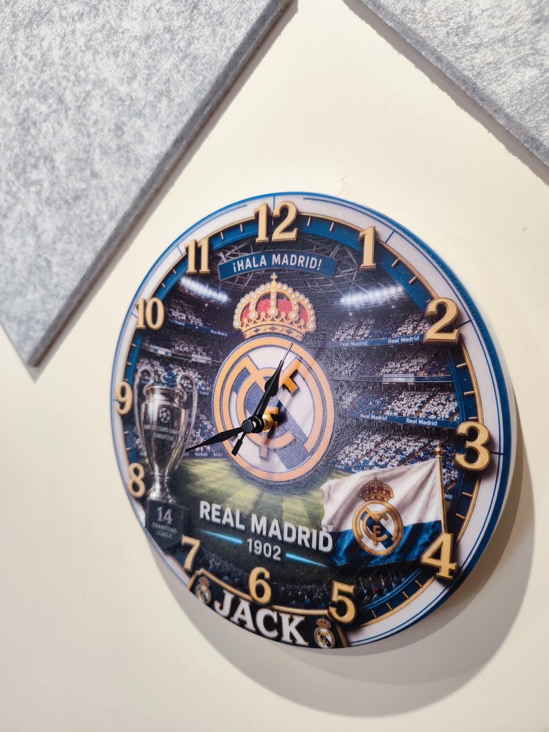 May include: A round wall clock featuring a Real Madrid theme with a stadium background, the club crest, a trophy, and a flag. The clock face displays the text "&iexcl;HALA MADRID!", "REAL MADRID 1902", and the name "JACK" at the bottom with gold numbers.
