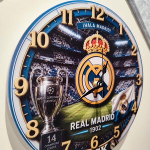 May include: A round wall clock featuring a Real Madrid theme with a stadium background, the club crest, a Champions League trophy, and the text "&iexcl;Hala Madrid!", "Real Madrid 1902", "14 Champions League", and "Jack" with gold-colored numbers.