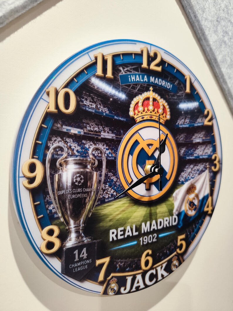 May include: A round wall clock featuring a Real Madrid theme with a stadium background, the club crest, a Champions League trophy, and the text "&iexcl;Hala Madrid!", "Real Madrid 1902", "14 Champions League", and "Jack" with gold-colored numbers.