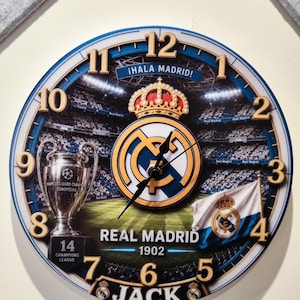 May include: A round wall clock featuring the Real Madrid logo, a stadium background, the Champions League trophy, and the text "&iexcl;HALA MADRID!", "REAL MADRID 1902", "14 CHAMPIONS LEAGUE", and "JACK". It has gold-colored numbers 1 to 12 and black hands.