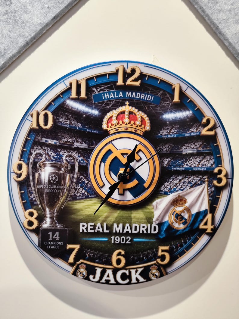 May include: A round wall clock featuring the Real Madrid logo, a stadium background, the Champions League trophy, and the text "&iexcl;HALA MADRID!", "REAL MADRID 1902", "14 CHAMPIONS LEAGUE", and "JACK". It has gold-colored numbers 1 to 12 and black hands.
