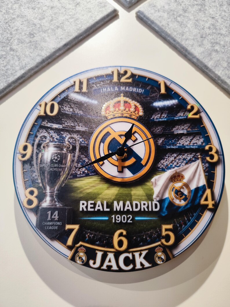 May include: A round wall clock featuring the Real Madrid logo, a stadium background, the Champions League trophy with the number 14, a team flag, and the text "&iexcl;HALA MADRID!", "REAL MADRID 1902", and "JACK" with black hour, minute, and second hands.