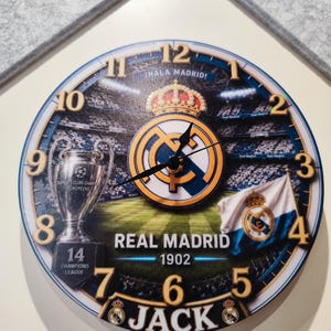 May include: A round wall clock featuring the Real Madrid logo, a stadium background, the Champions League trophy with the number 14, a team flag, and the text "&iexcl;HALA MADRID!", "REAL MADRID 1902", and "JACK" with black hour, minute, and second hands.