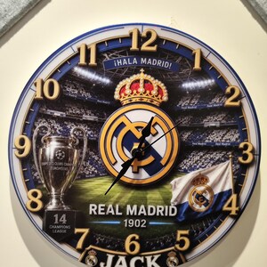May include: A round wall clock featuring a Real Madrid soccer stadium background, the team logo with a crown, a Champions League trophy, and a flag. The clock face displays the text &iexcl;HALA MADRID!, REAL MADRID 1902, 14 CHAMPIONS LEAGUE, and JACK.